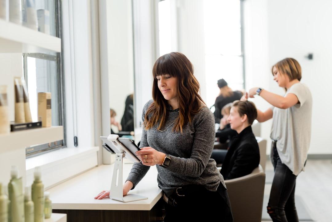 Professional woman managing booking system at salon reception desk with stylist working with client in background