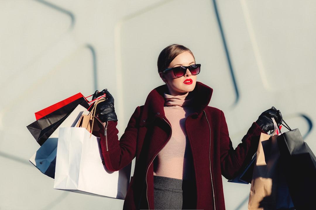 Stylish woman in burgundy coat and sunglasses holding colorful shopping bags against white wall
