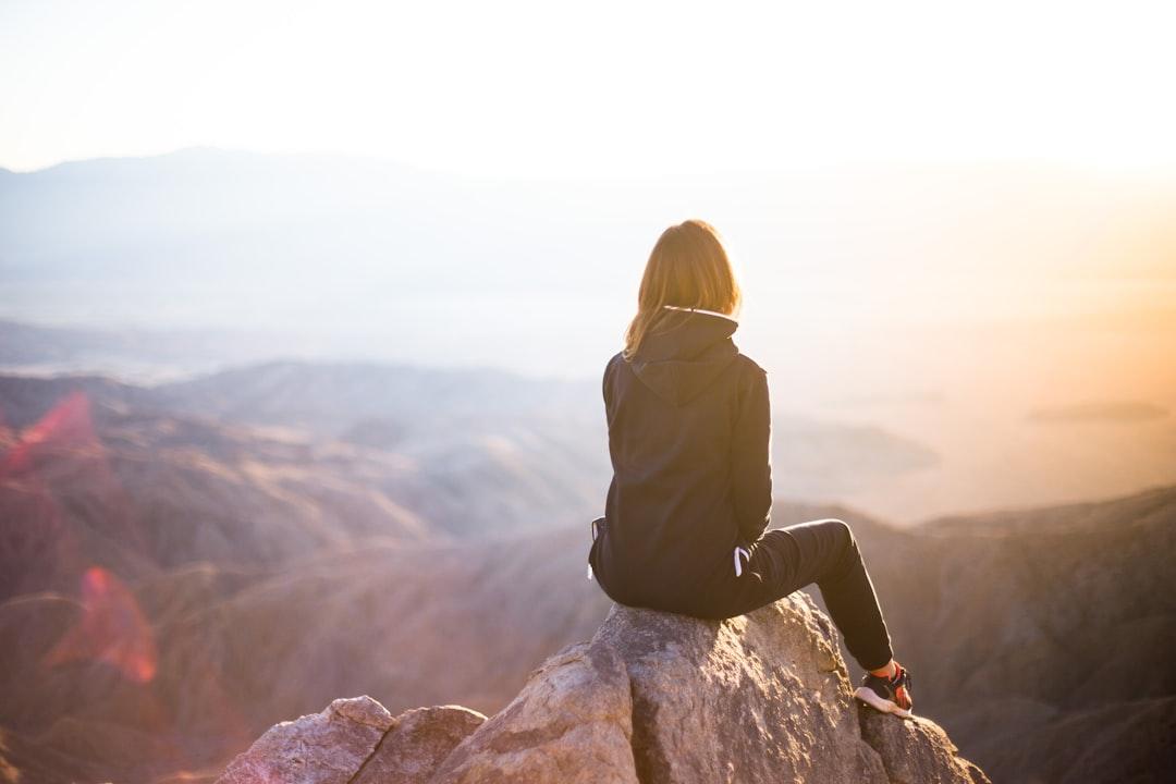 Woman sitting on mountain cliff overlooking valley at sunrise, representing personal growth and life coaching transformation.