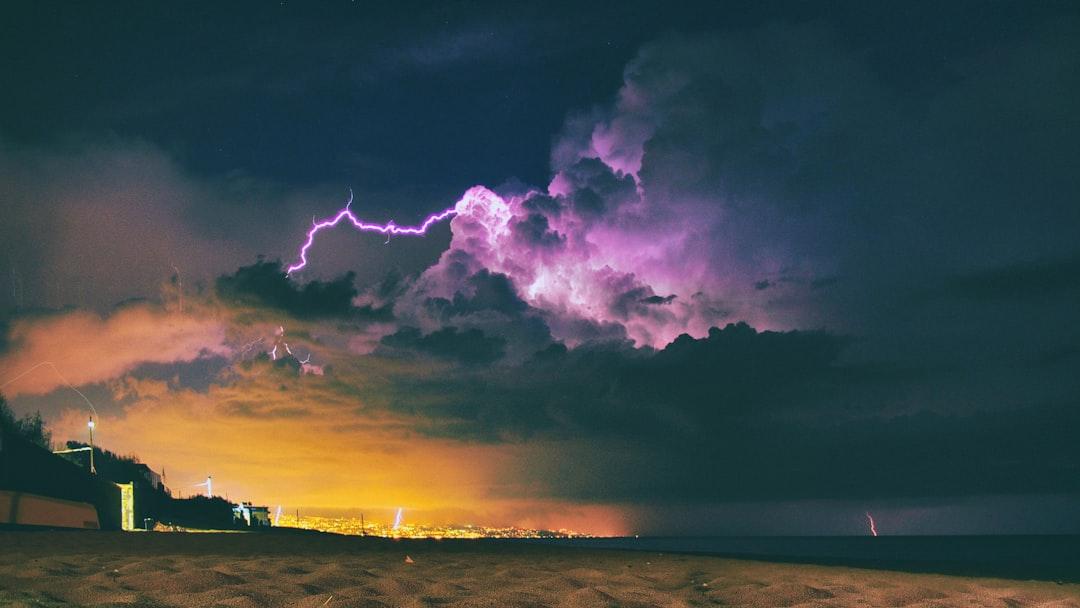 Dramatic nighttime thunderstorm with purple lightning and golden city lights illuminating coastal landscape below