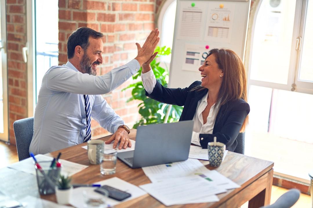 Two professionals high-fiving at desk with laptop and business documents in bright office space