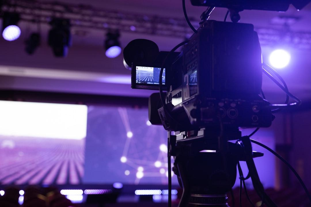 Professional video camera filming with purple stage lighting and perspective grid display on monitor